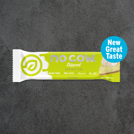 A single No Cow Dipped protein bar in lime key lime pie flavor, shown horizontally on a dark textured background with a blue circular badge that reads 'New Great Taste'.