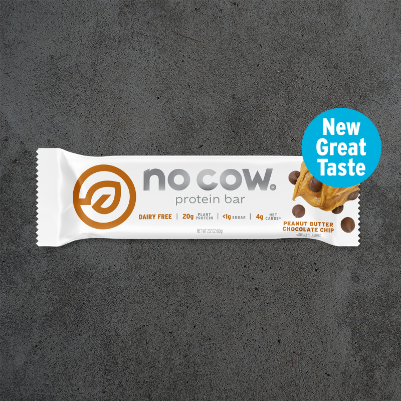 A single No Cow protein bar in a white wrapper with orange and gray branding, placed on a dark textured surface. A blue circular badge on the right reads 'New Great Taste' and includes a small peanut butter chocolate chip illustration.