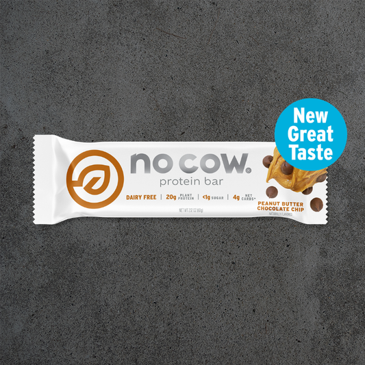 A single No Cow protein bar in a white wrapper with orange and gray branding, placed on a dark textured surface. A blue circular badge on the right reads 'New Great Taste' and includes a small peanut butter chocolate chip illustration.